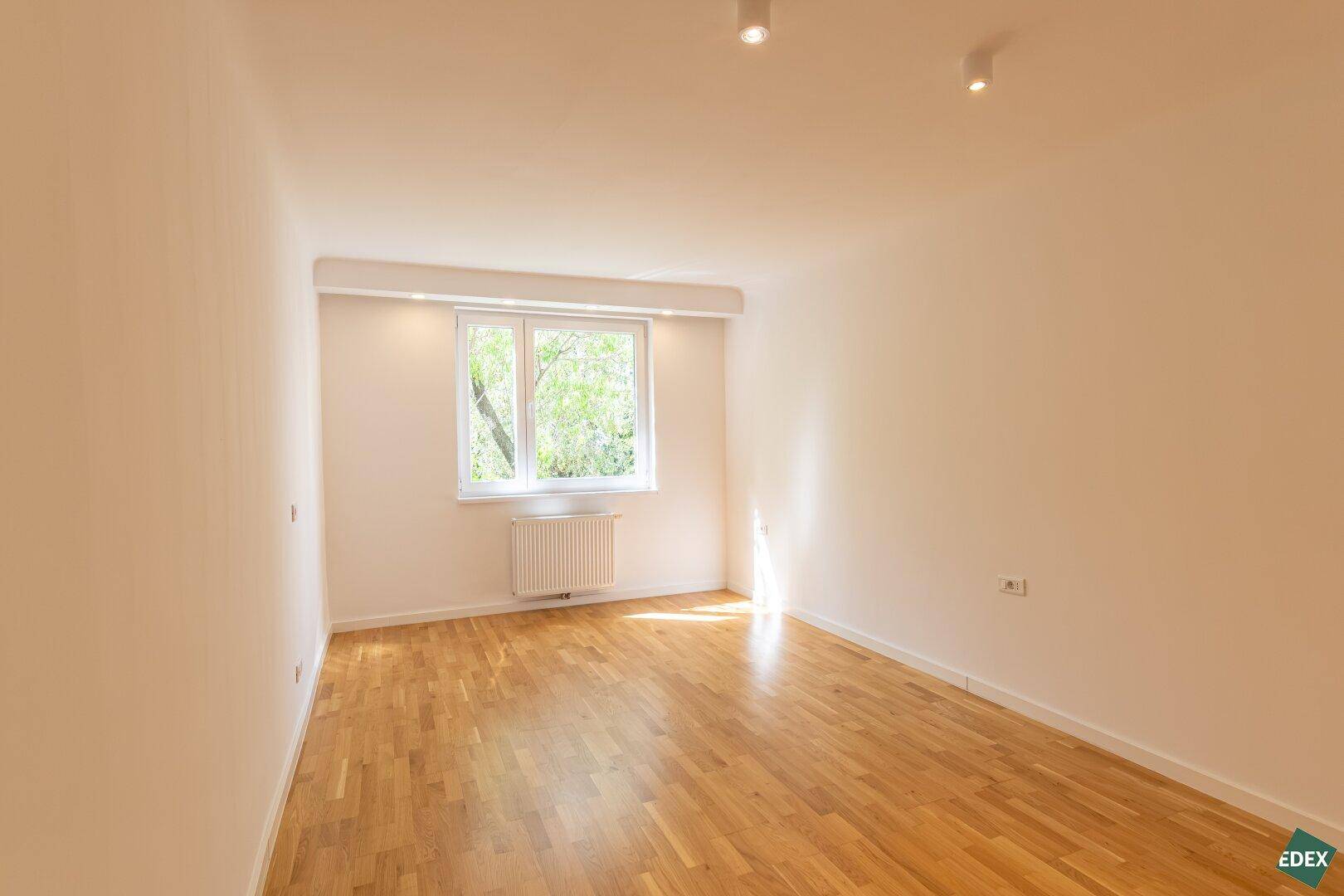 Helles Schlafzimmer mit Parkettboden und großem Fenster, das viel Tageslicht hereinlässt.
