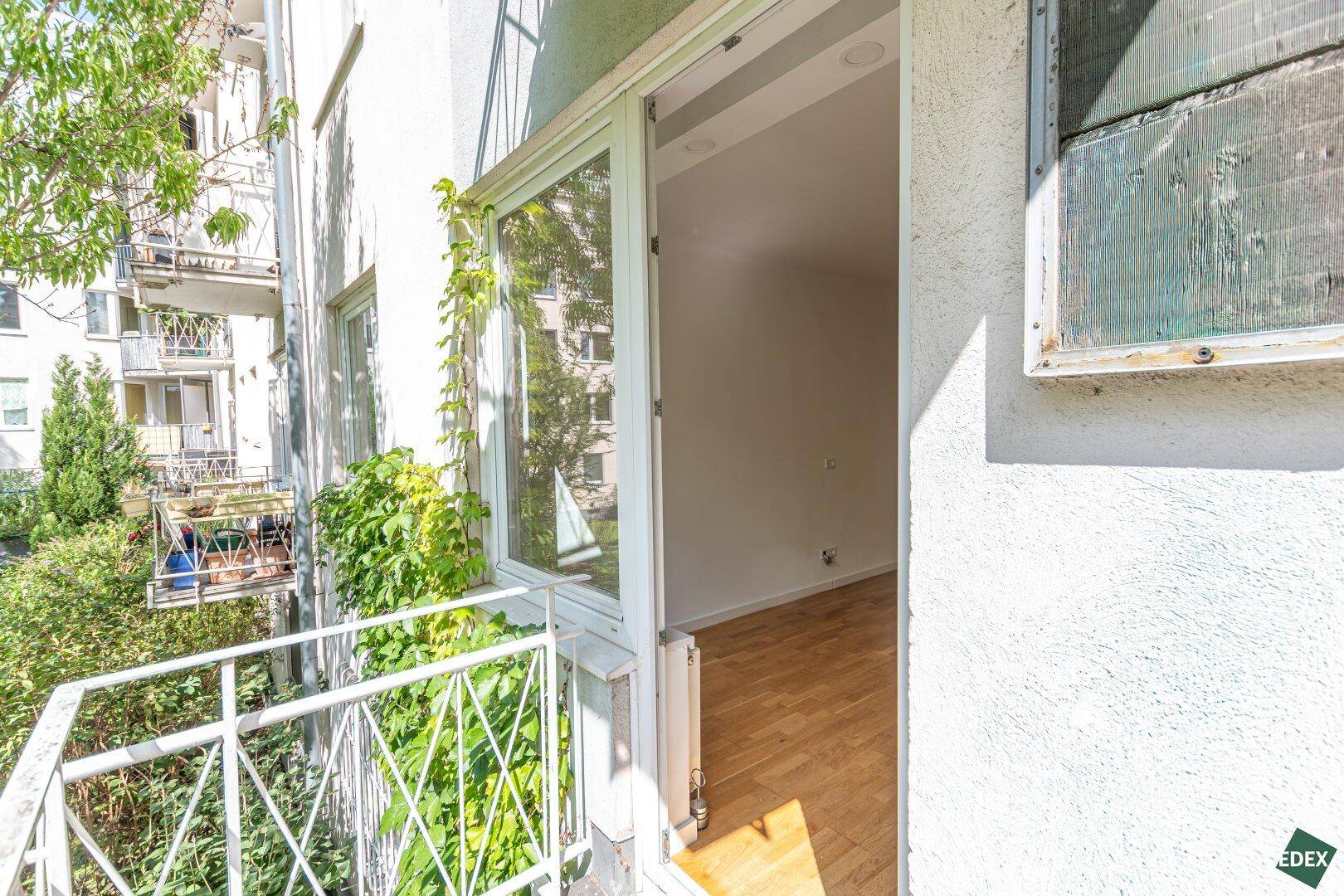 Blick vom Balkon in einen hellen Raum mit Parkettboden und viel Grün.