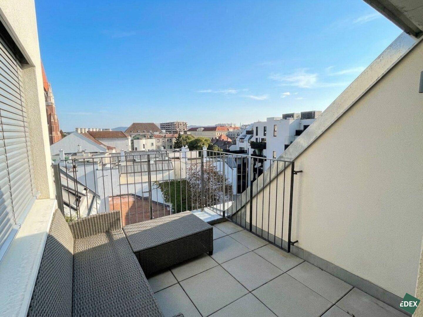 Sonniger Balkon mit Sitzgelegenheiten und Blick über die Dächer der Stadt.