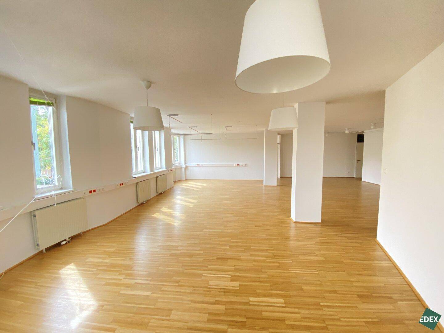 Großer, heller Büroraum mit Holzparkettboden, vielen Fenstern und modernen Hängelampen, ideal für offene Arbeitsbereiche.