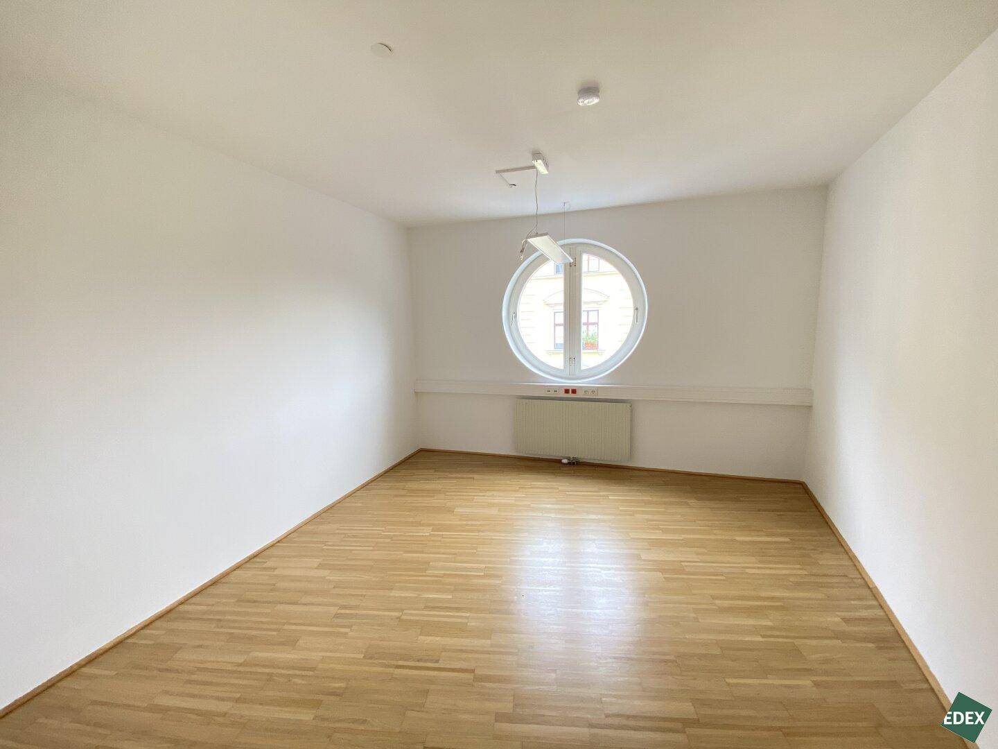 Heller Raum mit markantem Rundfenster, Holzparkettboden und weißen Wänden, vielseitig als Büro oder Arbeitszimmer nutzbar.