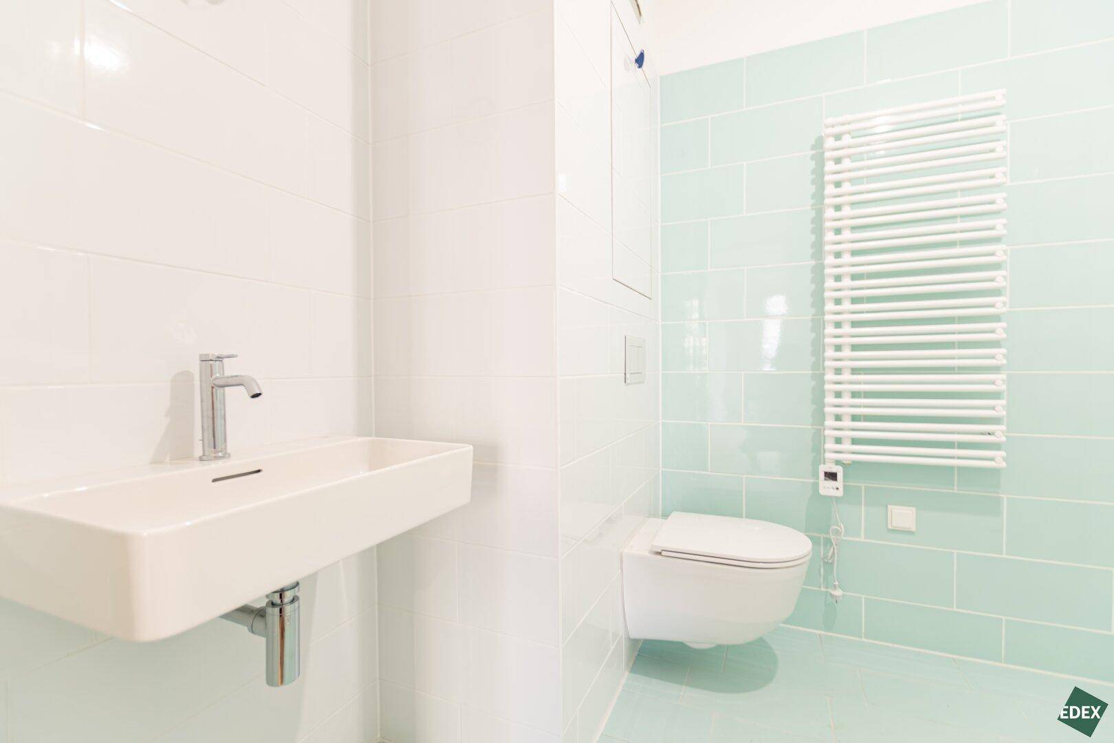 Modernes Badezimmer mit hellblauen Fliesen, wandhängendem WC, Waschbecken und Handtuchheizkörper.