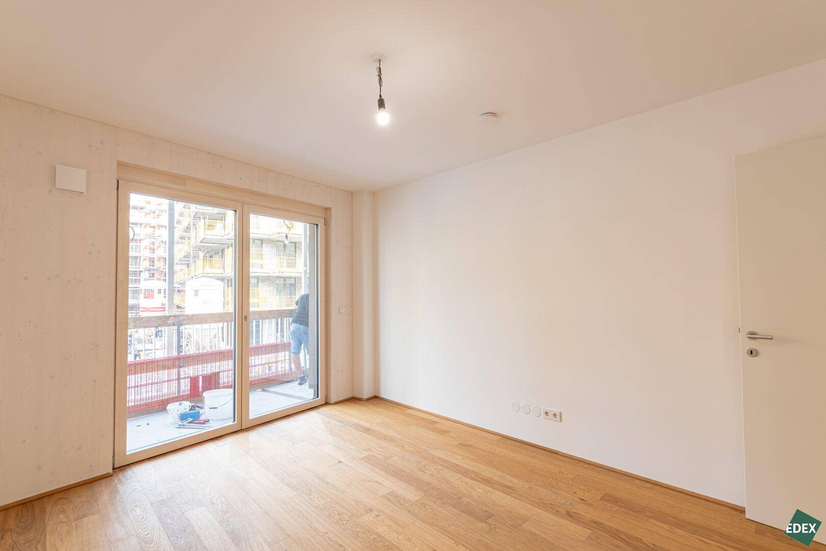 Helles Zimmer mit Holzboden und großem Fenster mit Zugang zu einem Außenbereich.