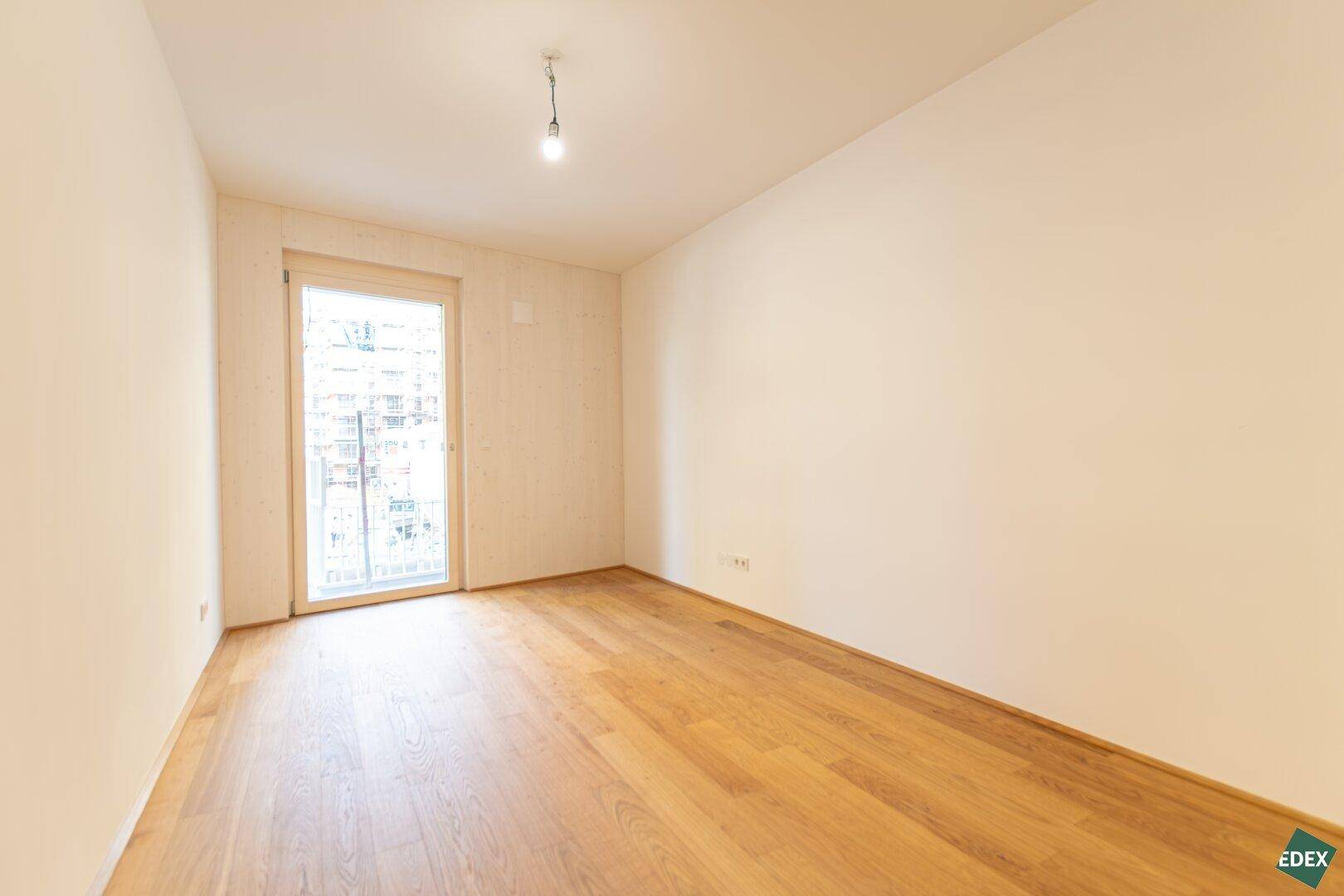 Leeres Zimmer mit Holzboden und Balkonzugang, bereit zur individuellen Gestaltung in einem Neubau.