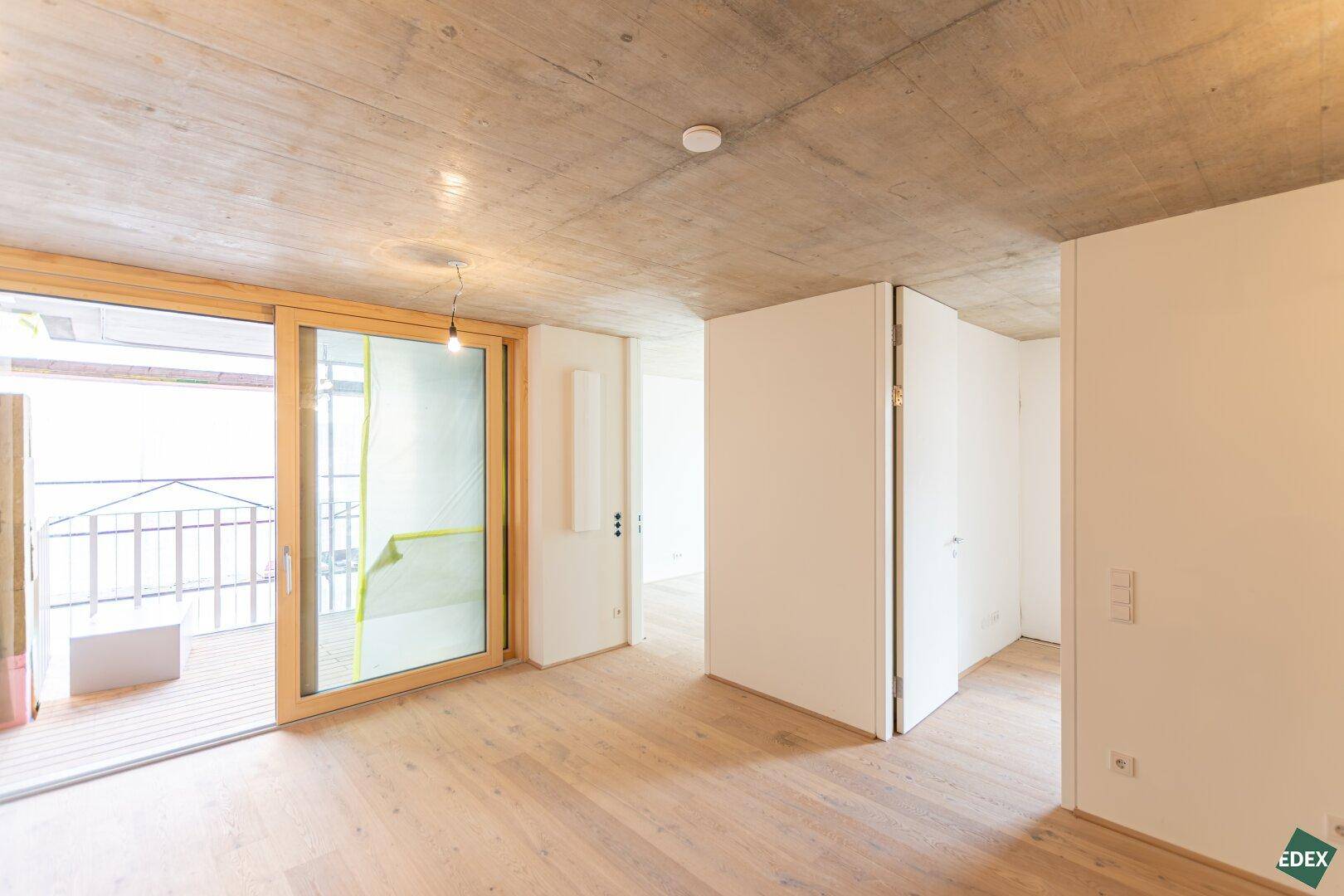 Lichtdurchfluteter Raum mit Schiebetür zum Balkon, Holzboden und moderner Betonoptikdecke.
