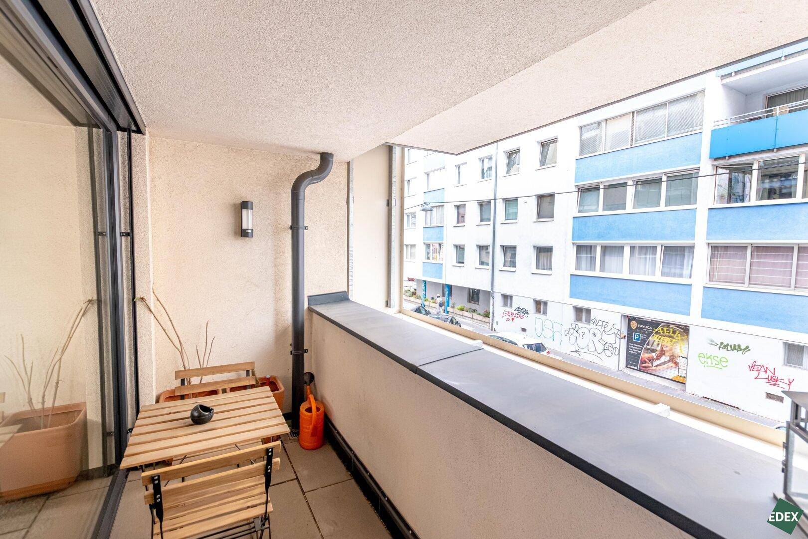 Schmaler Balkon mit Holztisch und Stühlen, bietet einen Blick auf die gegenüberliegenden Gebäude.