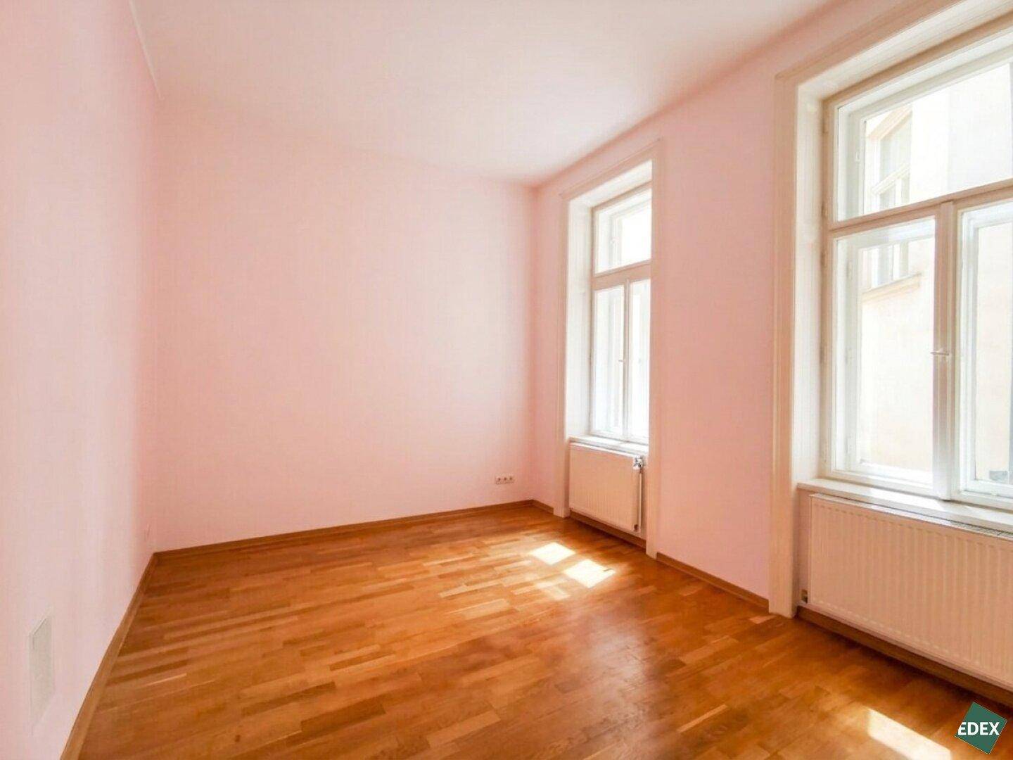 Gemütliches Zimmer mit hellrosa Wänden, Holzboden und zwei Fenstern, die den Raum erhellen.