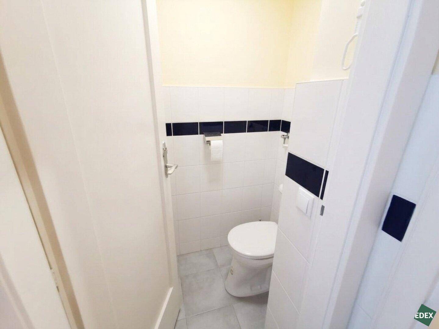 Kleines Gäste-WC mit weißen Fliesen, einer Toilette und einem Waschbecken, akzentuiert mit dunklen Fliesen.