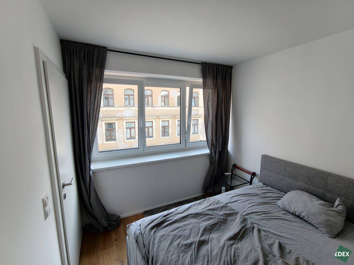 Schlafzimmer mit Holzboden, großem Fenster und Blick auf die umliegenden Gebäude.