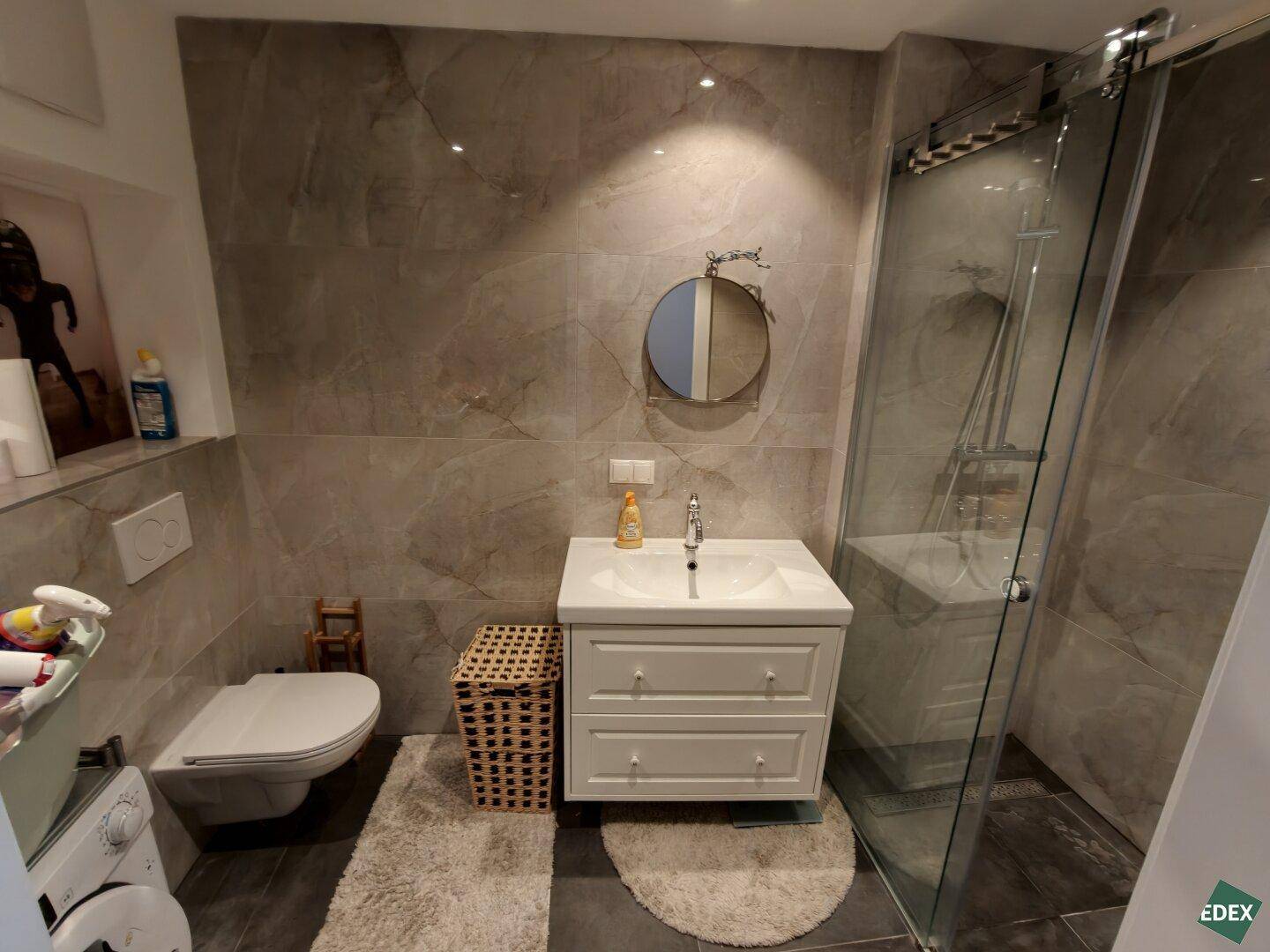 Modernes Badezimmer mit grauen Fliesen, Duschkabine und wandhängendem WC.