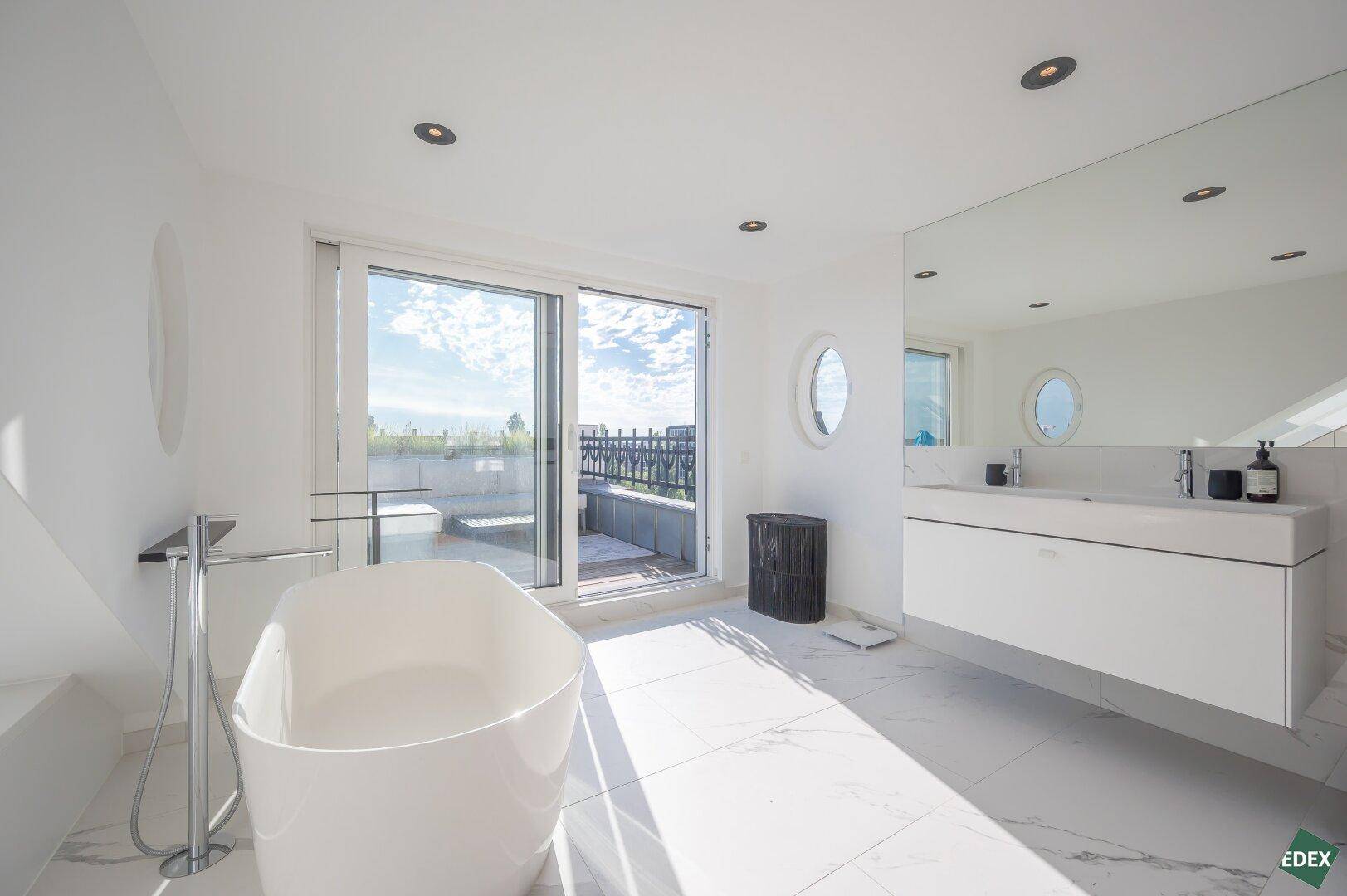 Geräumiges Badezimmer mit freistehender Badewanne, großem Spiegel und Zugang zu einer sonnigen Dachterrasse.