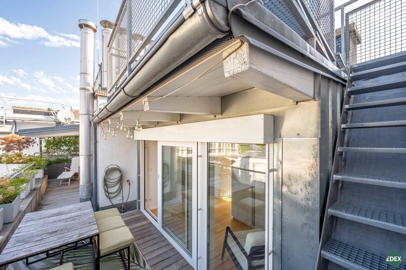 Dachterrasse mit Holzboden und Zugang zu den Innenräumen durch große Glasschiebetüren, umgeben von Metallgeländern.