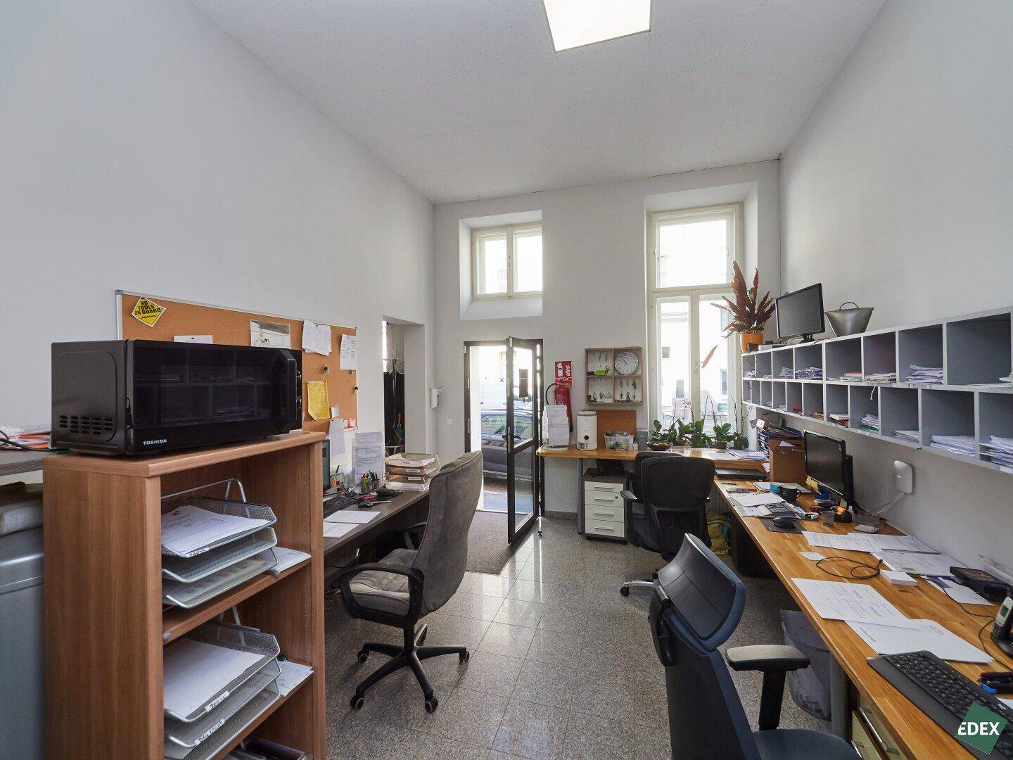 Helles Büro mit mehreren Arbeitsplätzen, Schreibtischen und Regalen, gut beleuchtet durch Fenster.