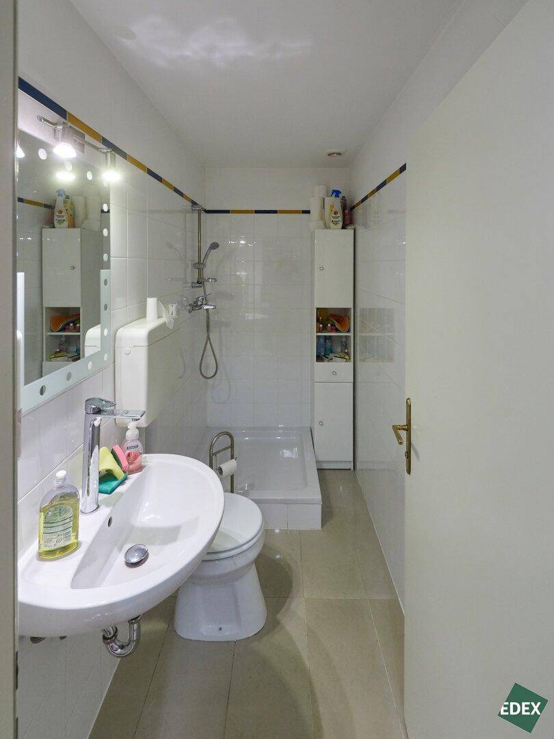 Schmaler Badezimmerbereich mit Waschbecken, Toilette und Duschkabine, hell gefliest.