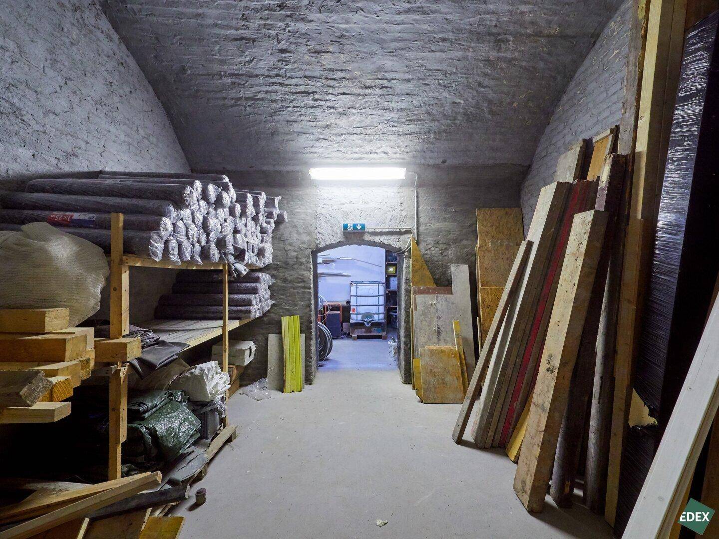 Langer, gewölbter Kellerraum, der als Lager für Materialien und Holzbretter genutzt wird.