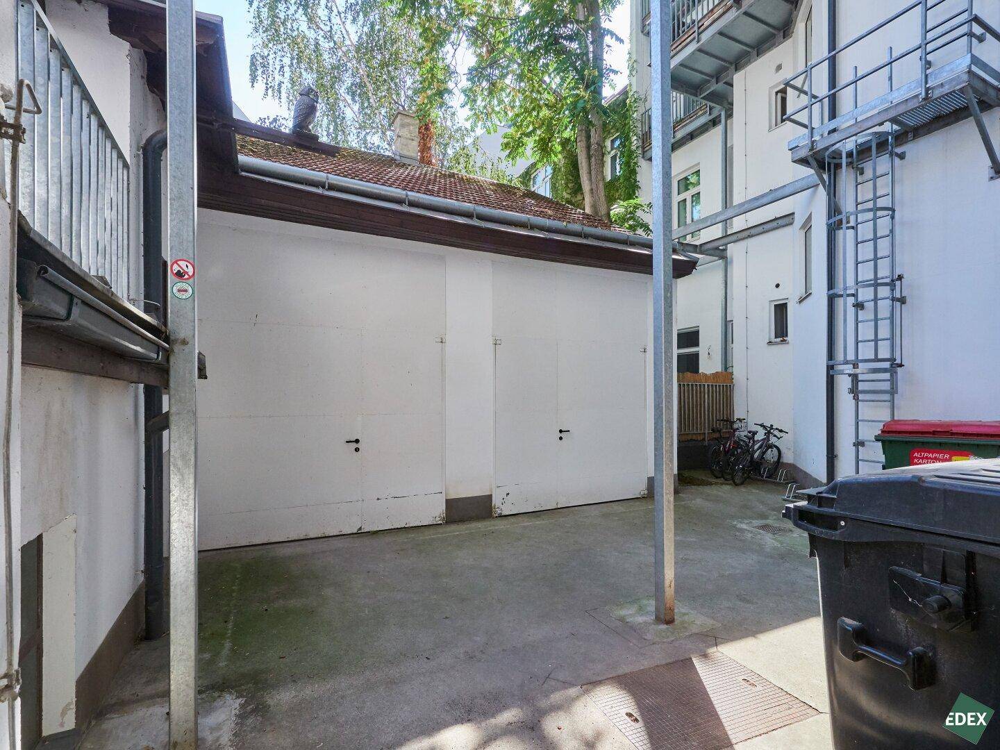 Hinterhof mit zwei Garagentoren und einem gepflasterten Bereich, umgeben von Gebäuden.
