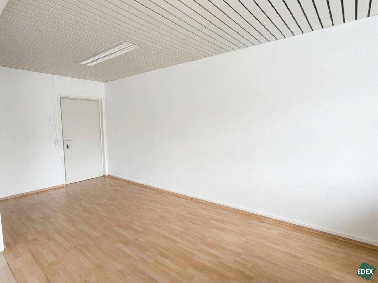 Leerer Büroraum mit weißer Tür, hellem Laminatboden und Deckenverkleidung.