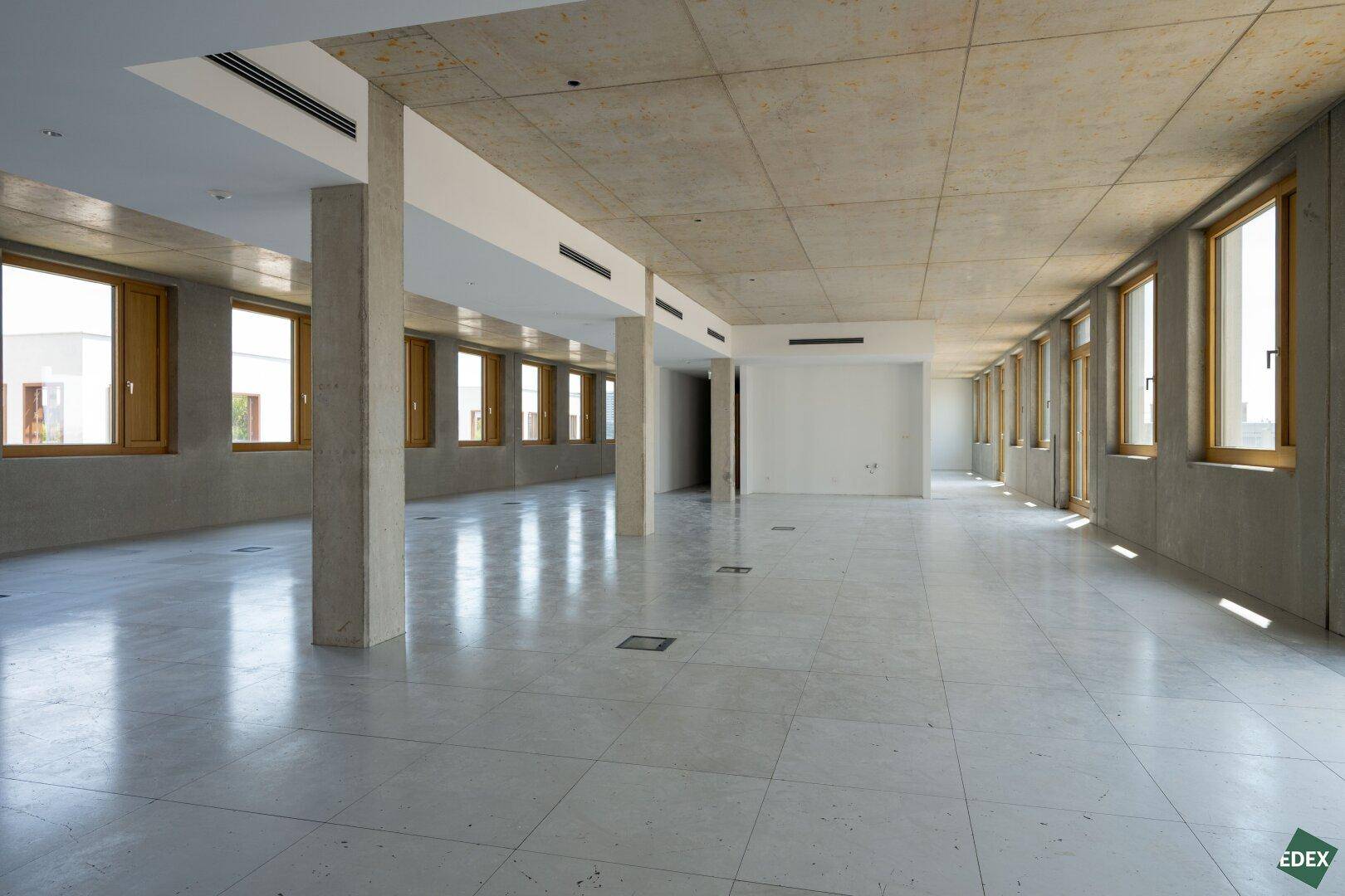 Weitläufiger Bürobereich mit Betonstützen, Fliesenboden und Lüftungsschlitzen in der Decke.