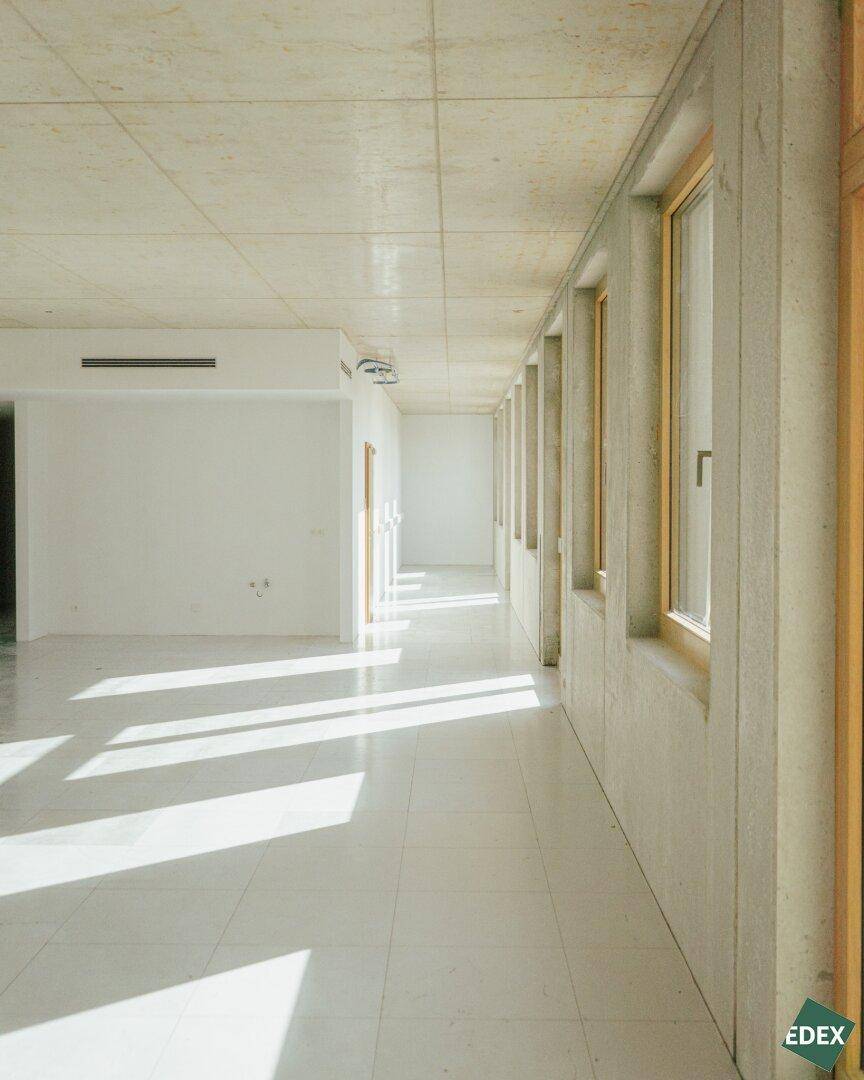 Heller Flur mit Fliesenboden, Betonwänden und Lüftungsschlitzen in der Decke, durchflutet von Sonnenlicht.