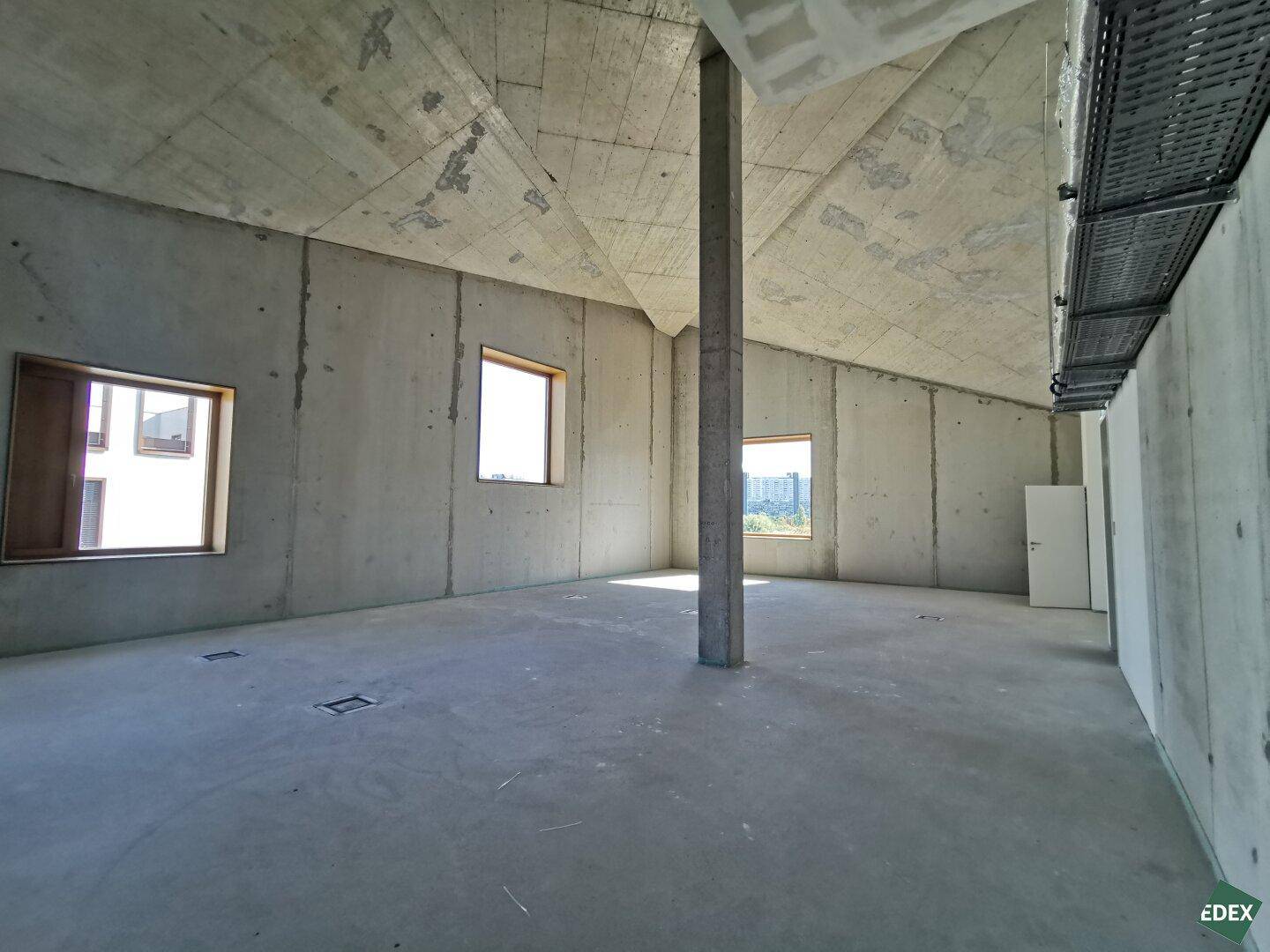 Dachgeschoss-Büroraum mit schrägen Betonwänden und -decke, einem Fenster und Betonboden.