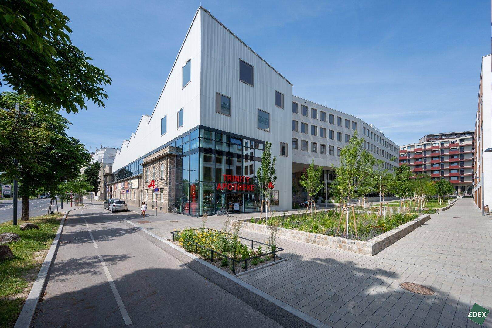 Ansicht eines modernen Gebäudekomplexes mit Apotheke im Erdgeschoss, umgeben von Bäumen und einem Gehweg.