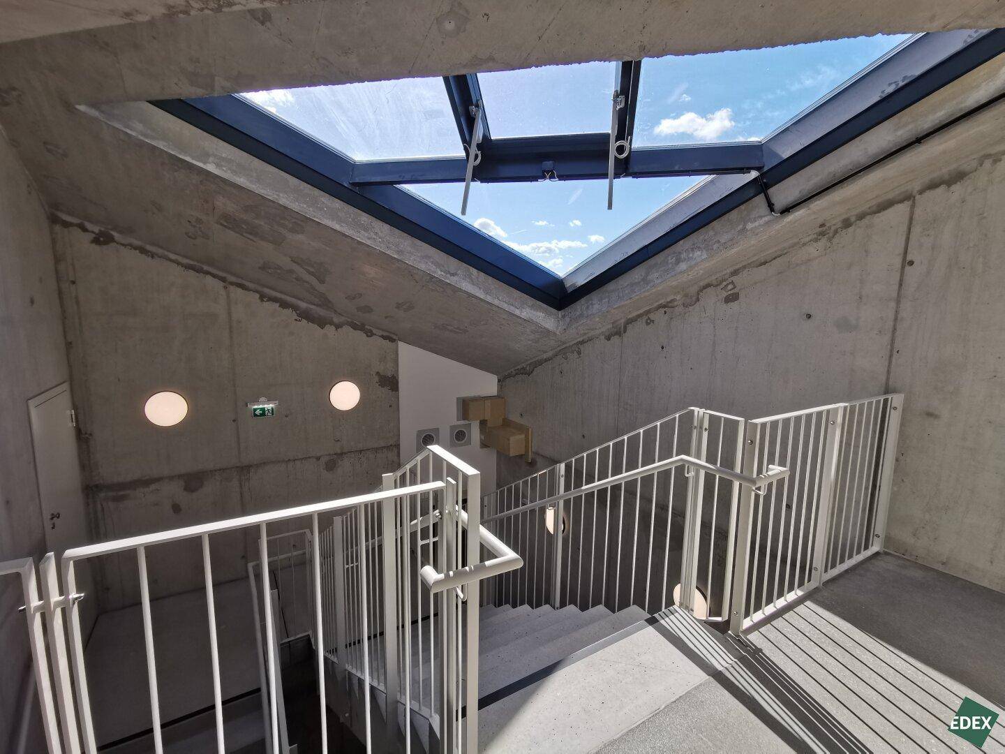 Treppenhaus mit Oberlicht, Betonwänden und Metallgeländer, das viel natürliches Licht hereinlässt.