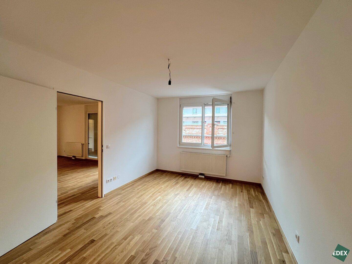 Geräumiges, unmöbliertes Zimmer mit hellem Parkettboden und Blick nach draußen.