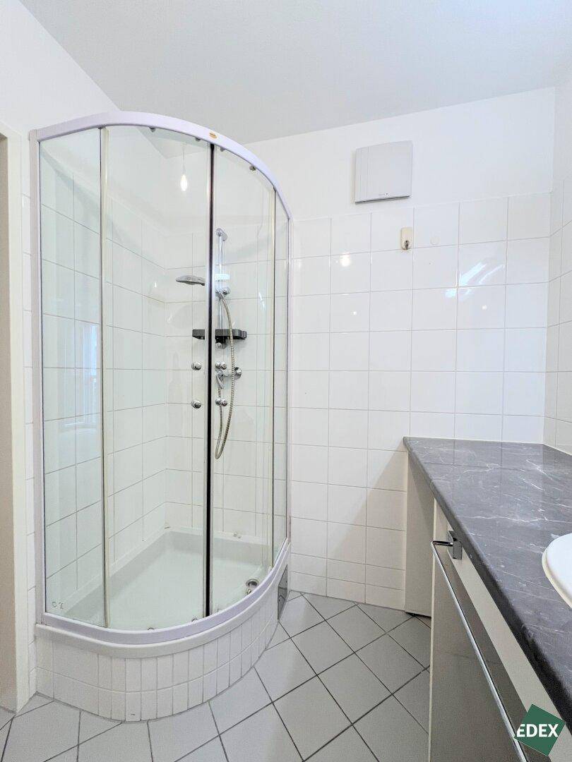 Modernes Badezimmer mit Duschkabine, weißen Fliesen und einem Waschtisch mit Ablagefläche.