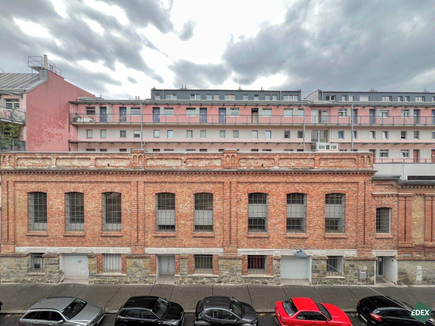 Blick auf die Fassade eines Backsteingebäudes mit darüberliegenden Wohnungen und parkenden Autos.