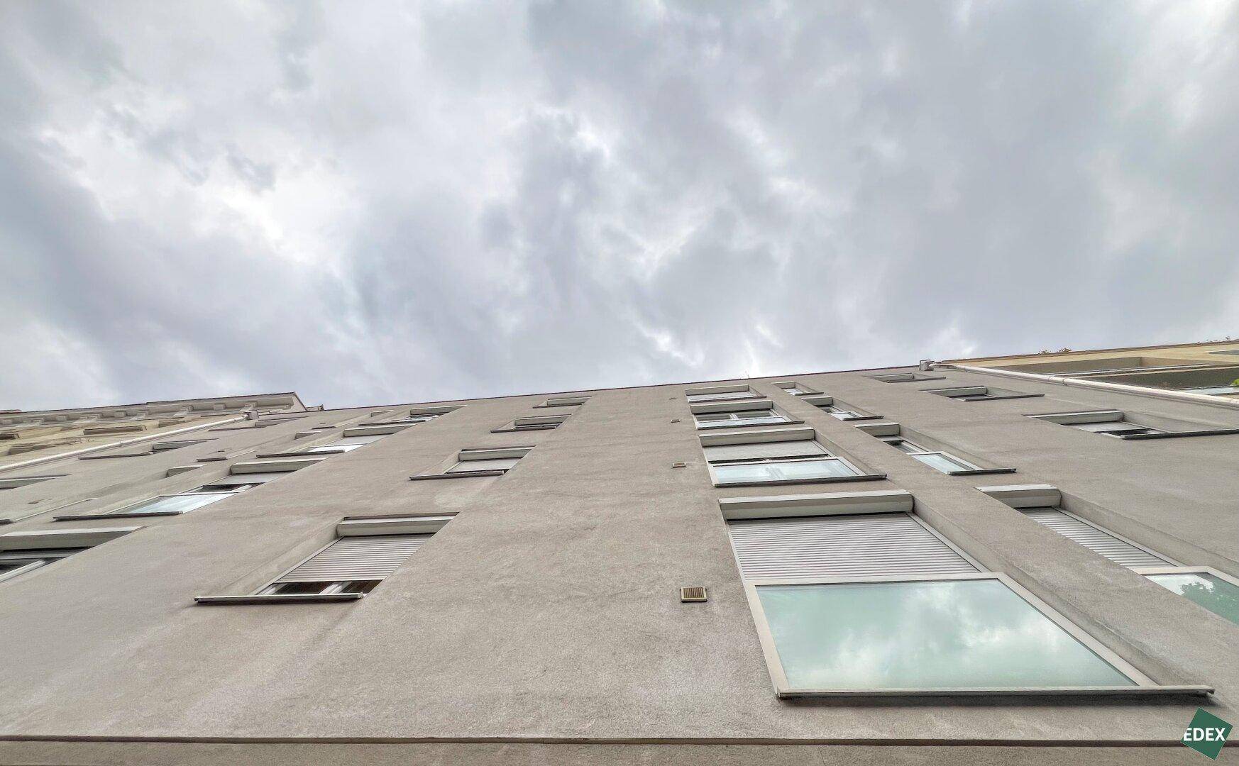 Ansicht einer modernen Gebäudefassade mit vielen Fenstern und Rollläden unter bewölktem Himmel.