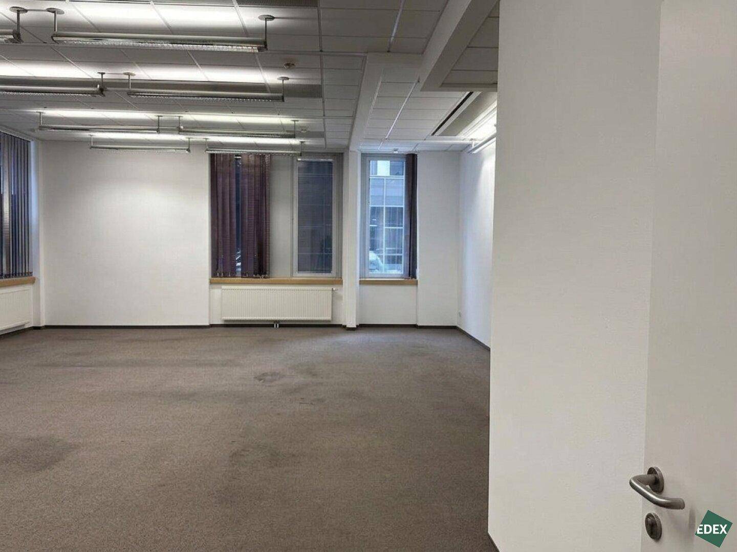 Leeres Büro mit Teppichboden, großen Fenstern und Jalousien, bereit zur Einrichtung.