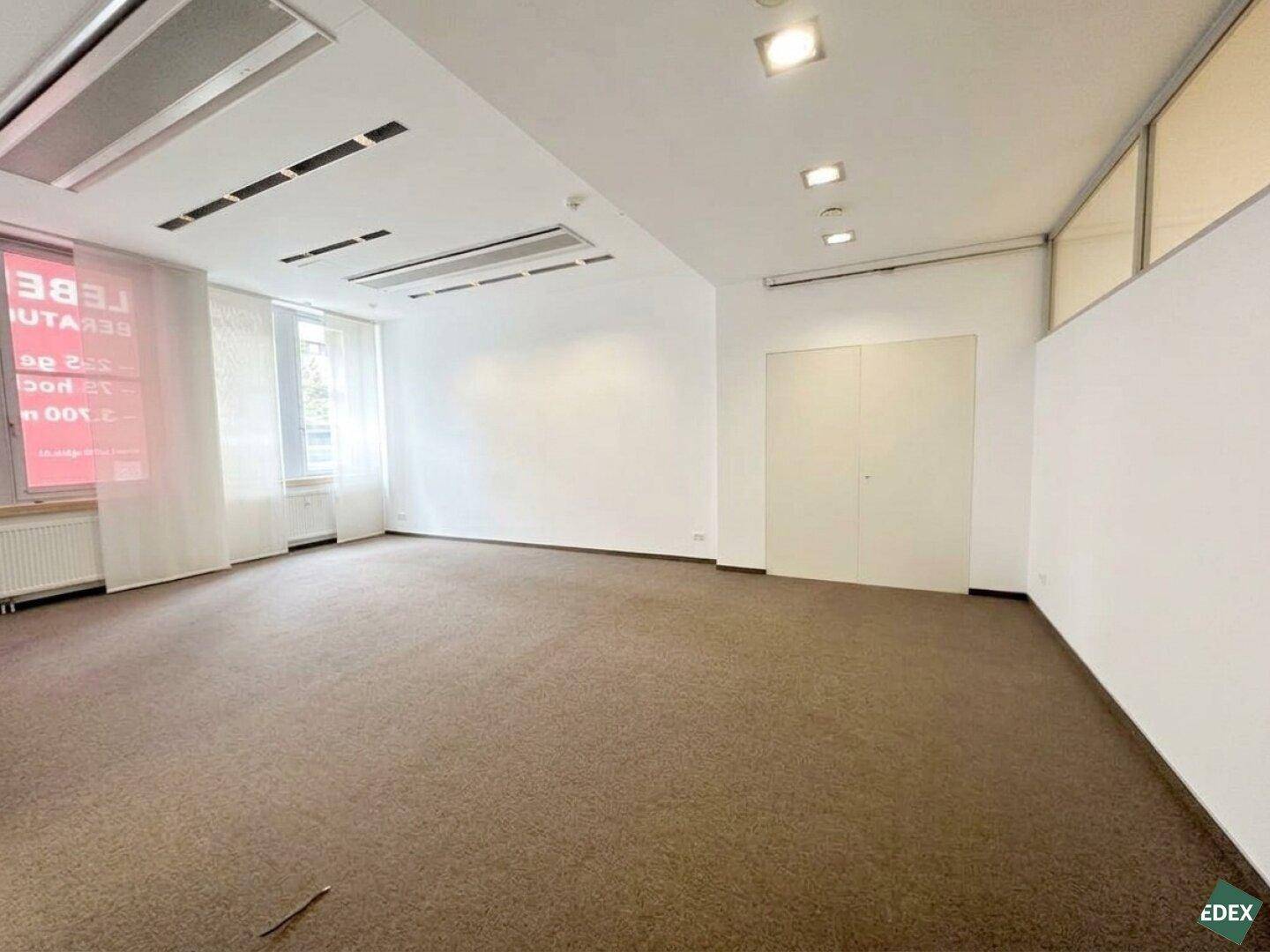 Geräumiges, leeres Büro mit Teppichboden, großen Fenstern und abgehängter Decke.