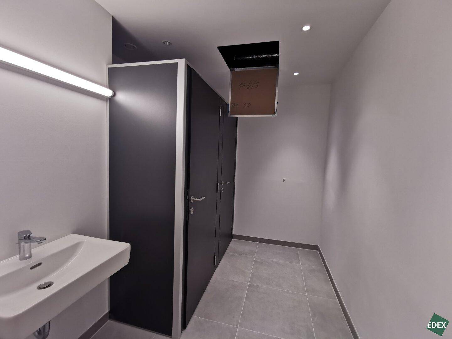 Modernes Badezimmer mit Waschbecken, hellen Wänden und dunklen Trennwänden, ausgestattet mit Abluftventilator.