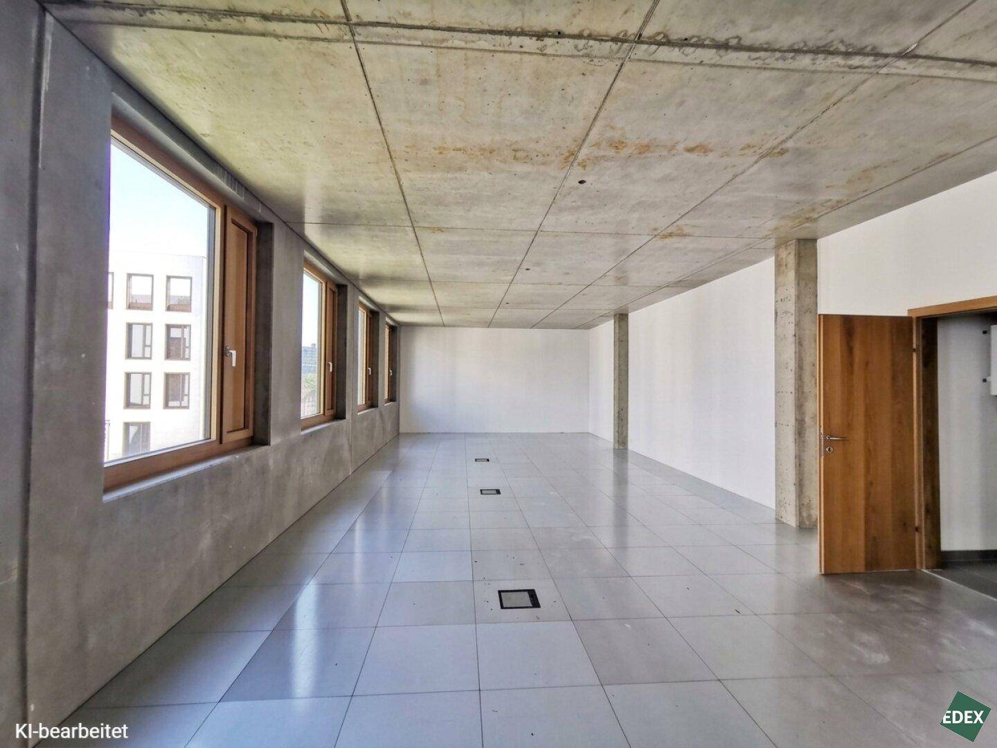Geräumiger, leerer Büroraum mit hohen Decken, großen Fenstern und Fliesenboden in einem Neubau.