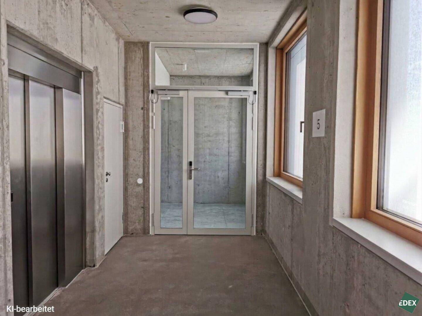 Moderner Flur mit Betonwänden, Aufzug und großen Fenstern mit Holzrahmen.