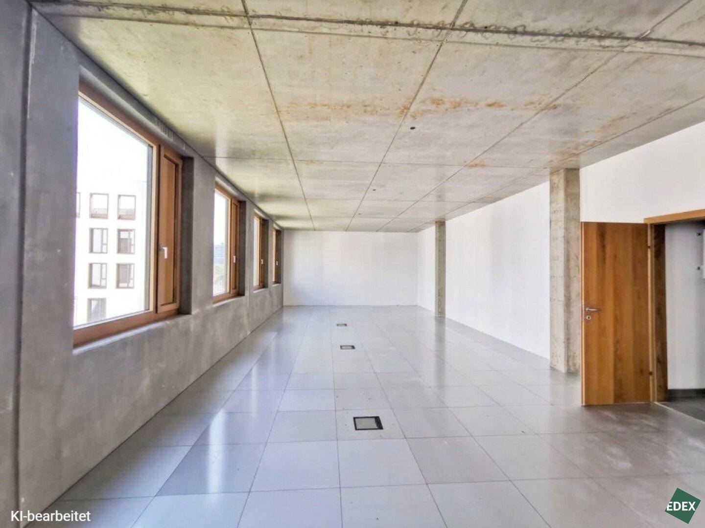 Geräumiger, leerer Büroraum mit Betonwänden, Fliesenboden und Holztür.