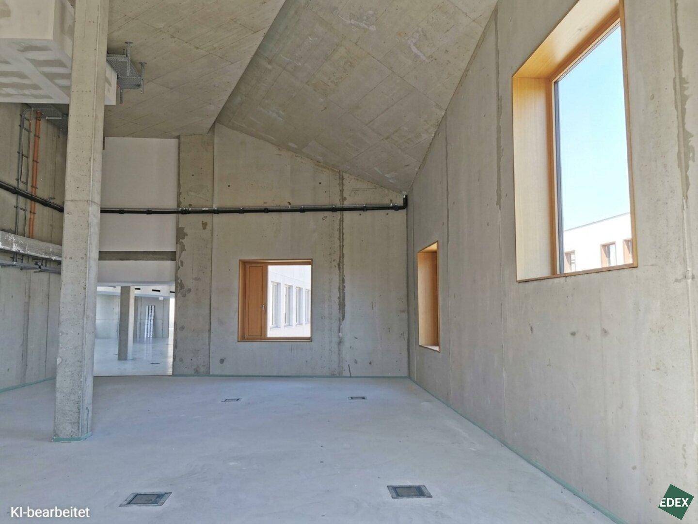 Geräumiger, unfertiger Innenraum mit schrägen Betondecken und großen Fenstern, ideal für Büronutzung.