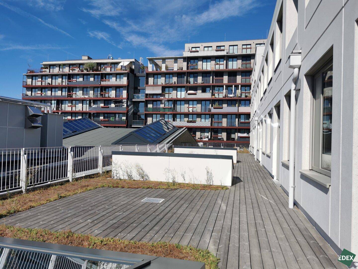 Weitläufige Dachterrasse mit Holzboden und Blick auf moderne Wohngebäude unter blauem Himmel mit Solaranlagen.