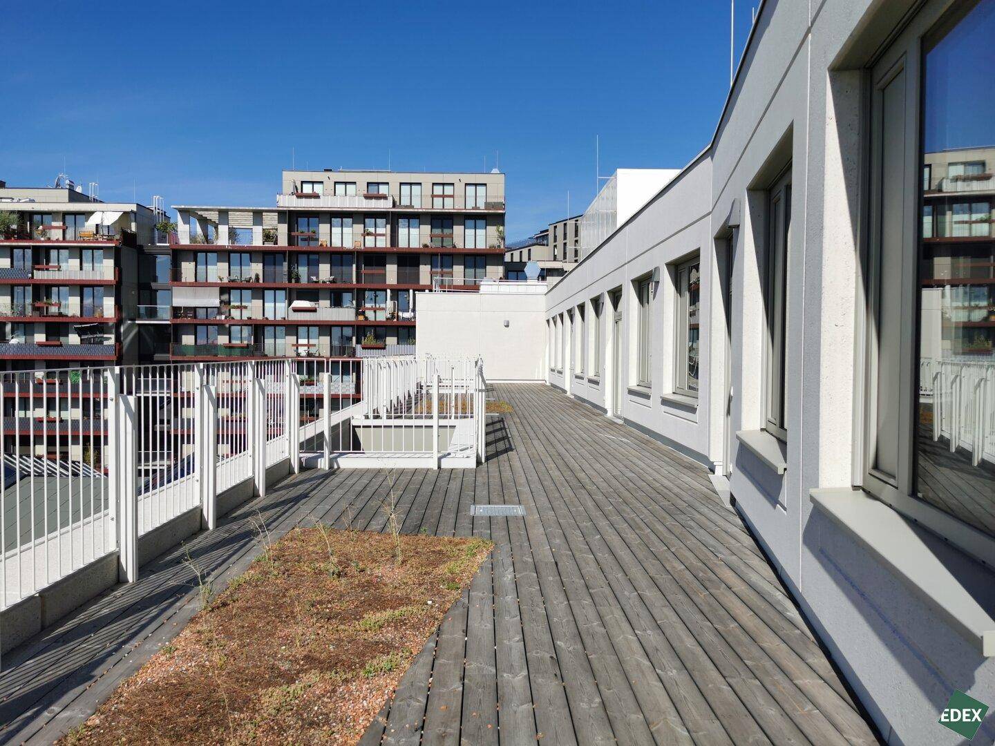 Weitläufige Dachterrasse mit Holzbelag und Metallgeländer, umgeben von modernen Gebäuden unter blauem Himmel.