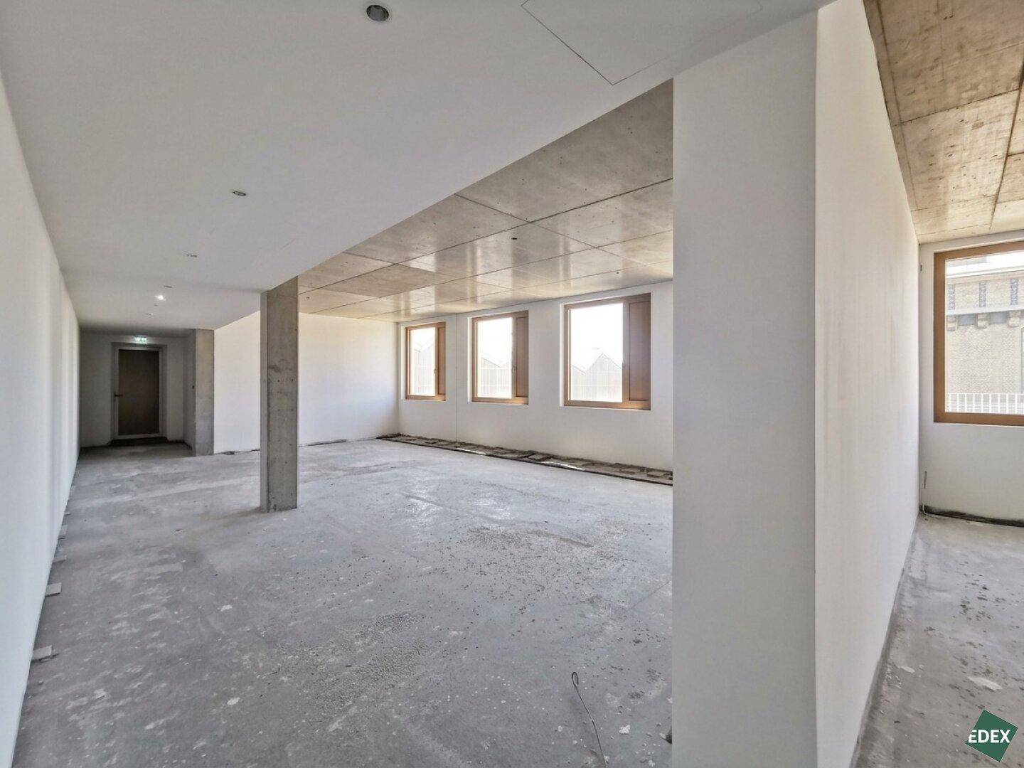 Großer, offener und unfertiger Innenraum mit Betonwänden und -decken sowie mehreren Fenstern für natürliches Licht.