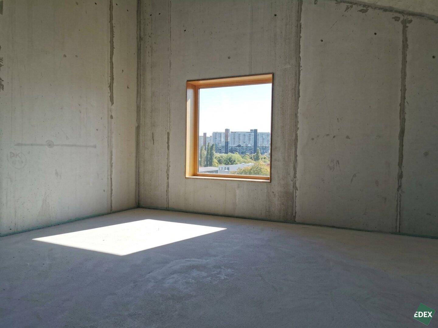 Unfertiger Raum mit Betonwänden und einem Fenster, das einen Blick auf die Stadt und viel Sonnenlicht bietet.
