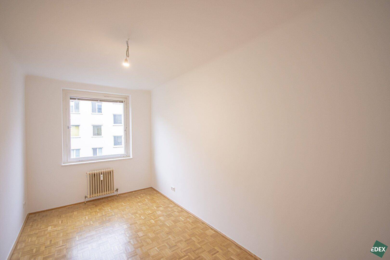 Kleines, helles Zimmer mit Parkettboden und einem Fenster mit Blick auf die Umgebung.