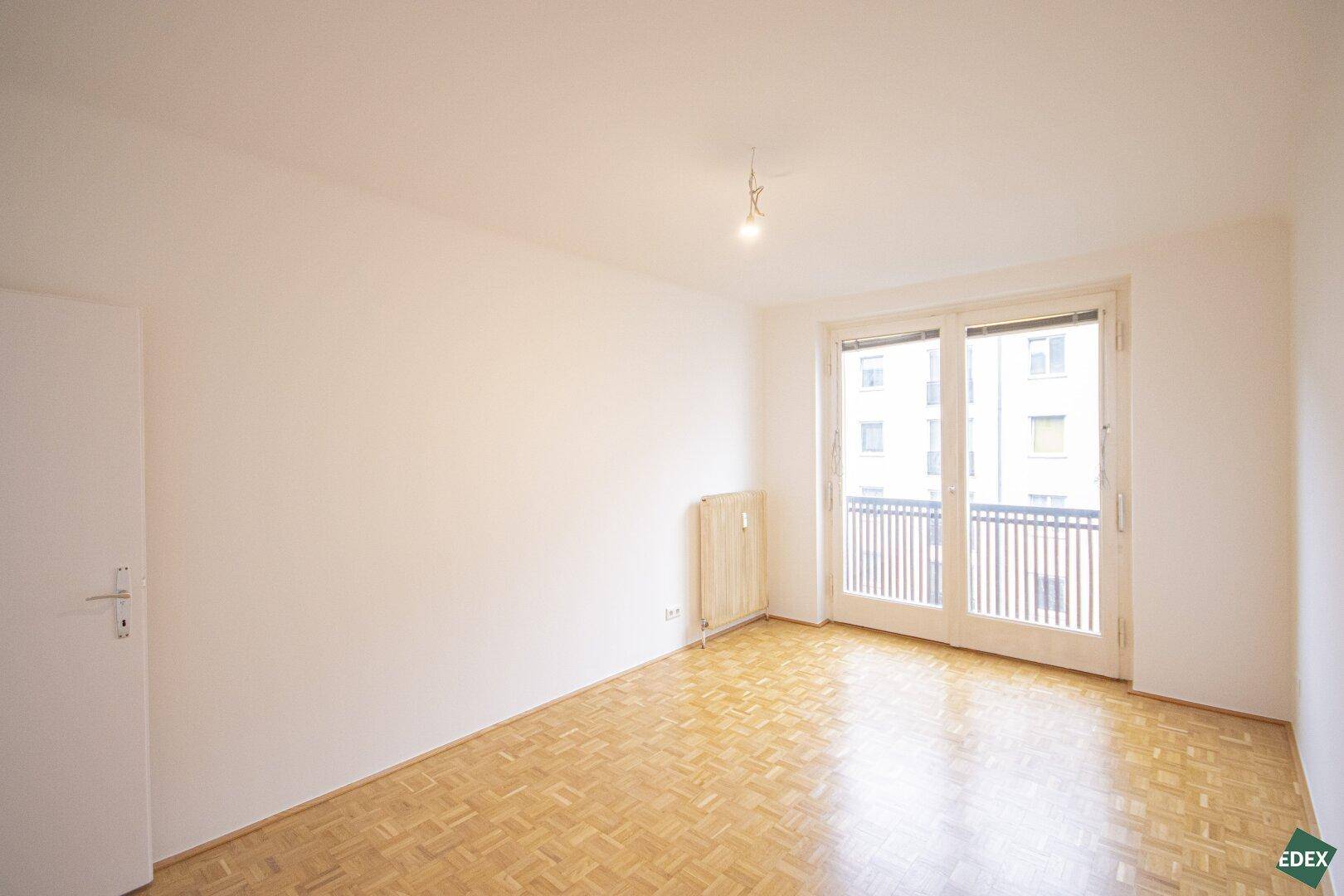 Geräumiges Wohnzimmer mit Parkettboden und großen Fenstern, die viel Licht hereinlassen.
