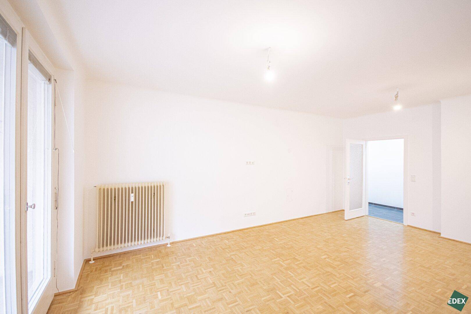 Heller Wohnbereich mit Parkettboden und großen Fenstern, die viel Tageslicht hereinlassen.