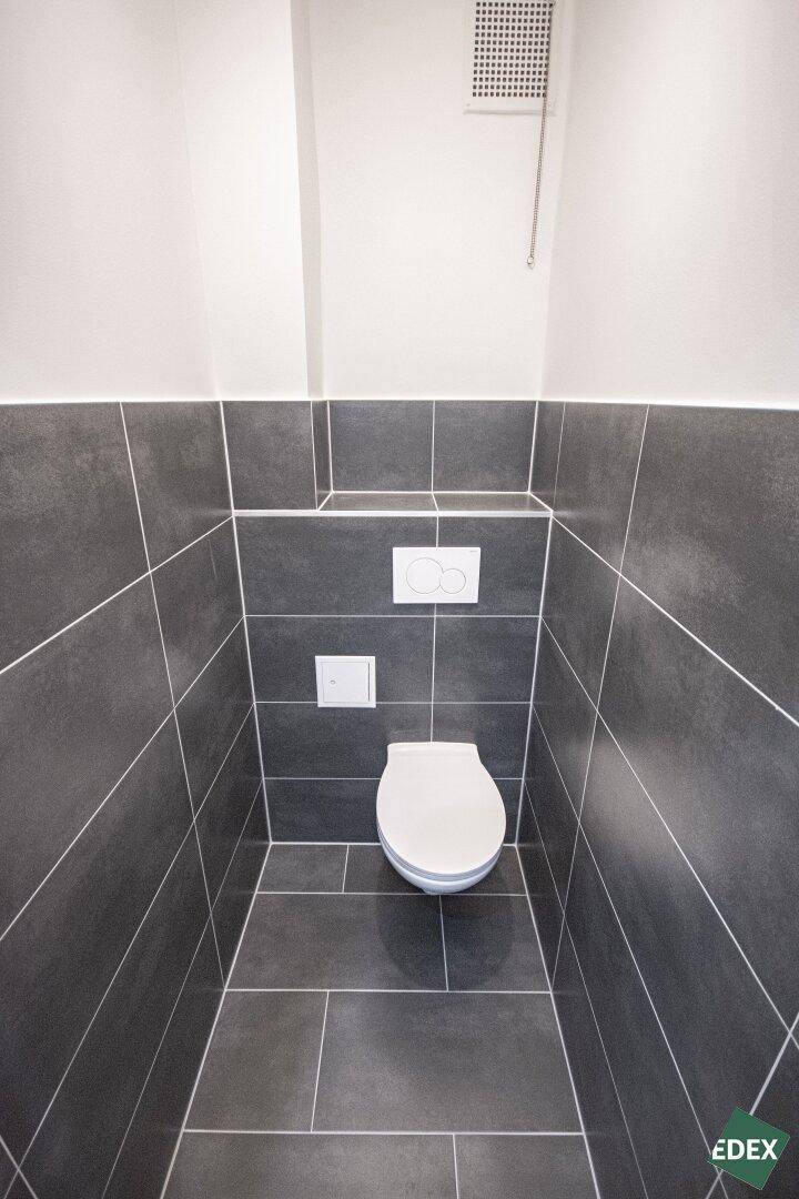 Separate Toilette mit Wand-WC und dunklen Fliesen an Wänden und Boden.