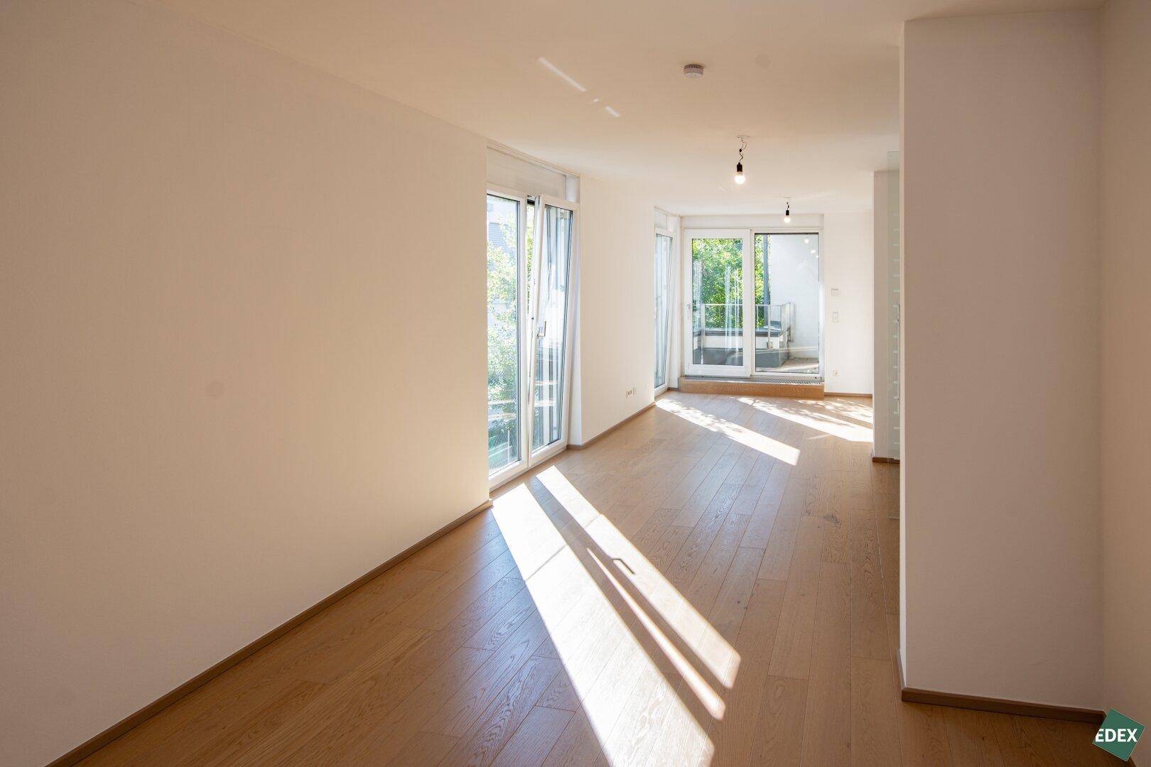 Lichtdurchfluteter Wohnraum mit hellem Holzboden und großen Fenstern, die den Raum erhellen und eine angenehme Atmosphäre schaffen.
