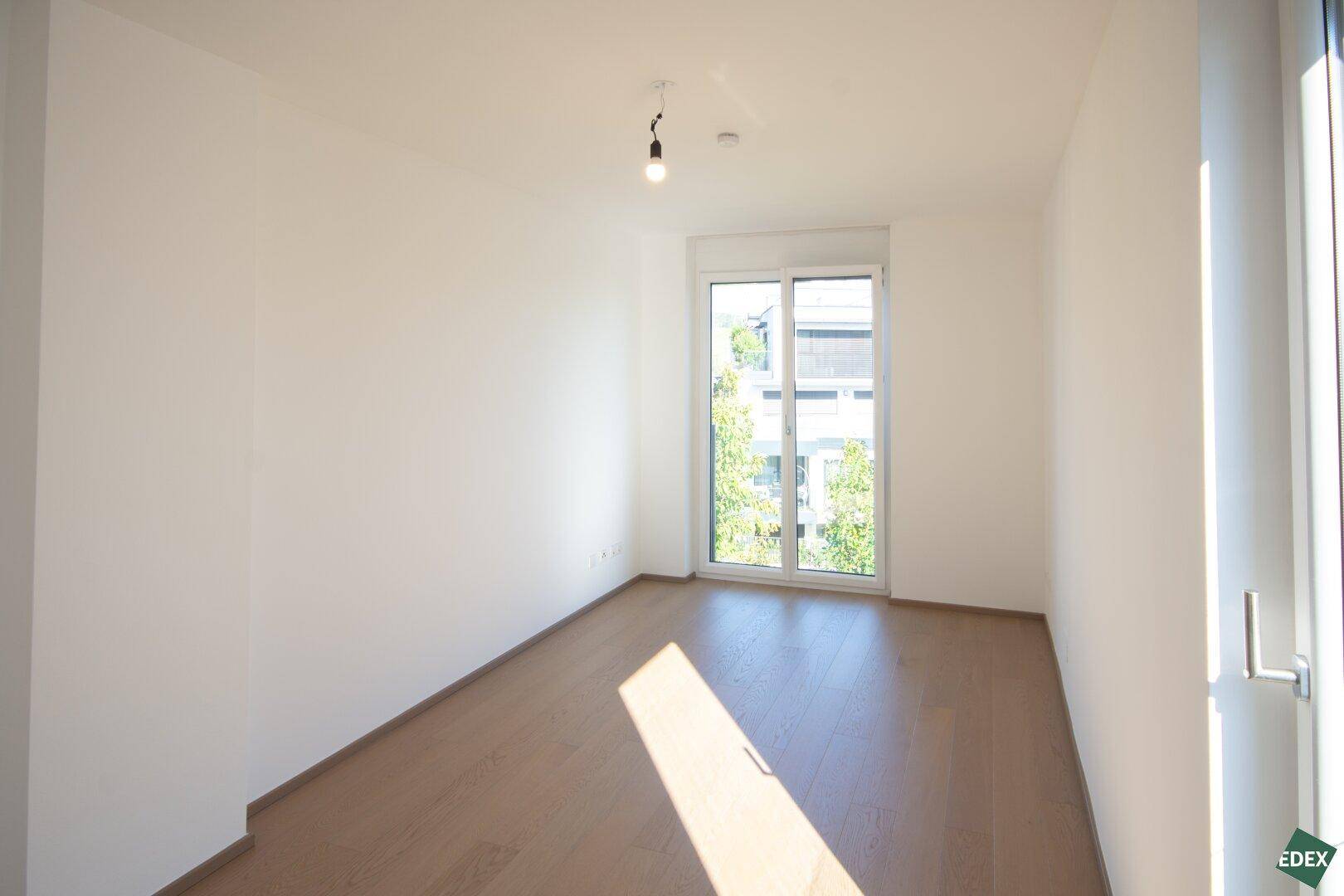 Helles Zimmer mit Holzboden und großem Fenster, das viel natürliches Licht spendet und eine freundliche Atmosphäre schafft.