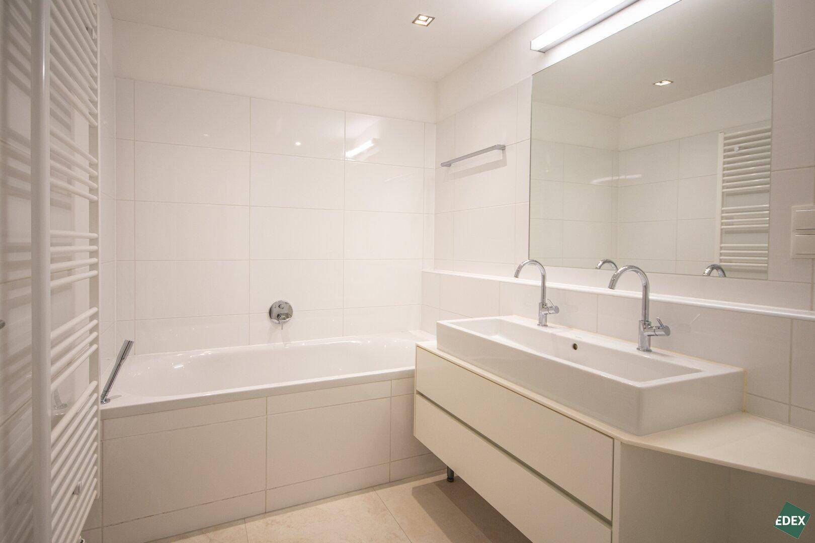 Modernes Badezimmer mit Badewanne, Doppelwaschbecken und weiß gefliesten Wänden, inklusive Handtuchheizkörper.
