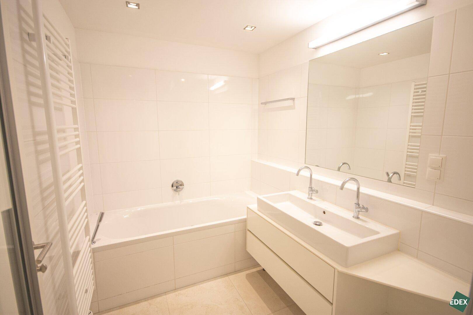 Helles Badezimmer mit Badewanne, Doppelwaschbecken und großem Spiegel, alles in Weiß gehalten und gut beleuchtet.