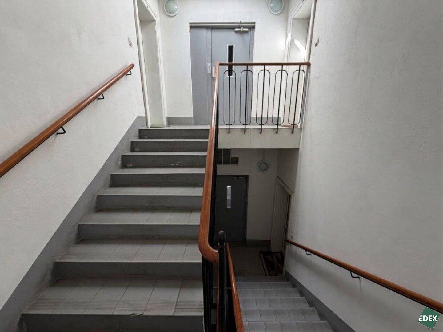Treppenhaus mit grauen Fliesenstufen, Holzgeländer und einem Aufzug im Hintergrund des Gebäudes.